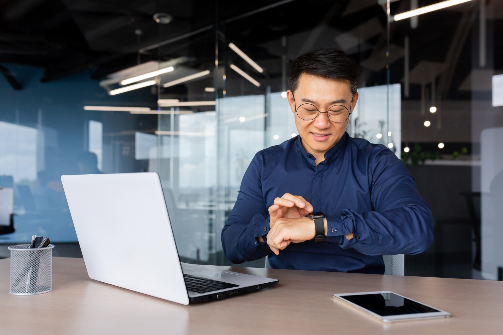 Successful asian businessman use smart watch at workplace, man chooses apps and smile happy with apps, employee inside office use laptop at work