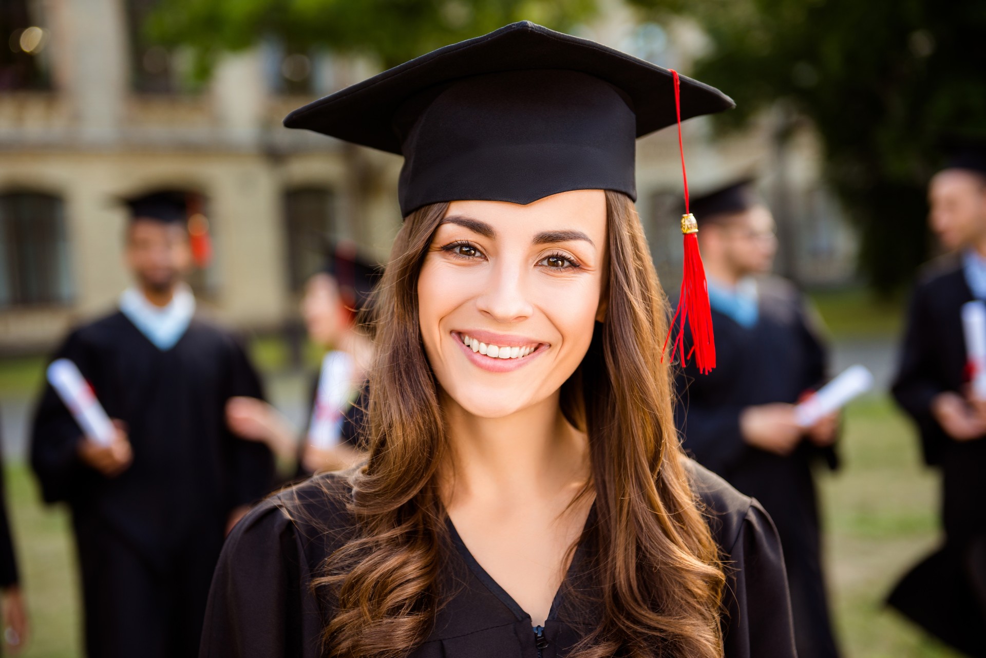 Happy cute brunette caucasian grad girl is smiling, her class mates are behind. She is in a black mortar board, with red tassel, in gown, with nice brown curly hair. Master degree! Success!